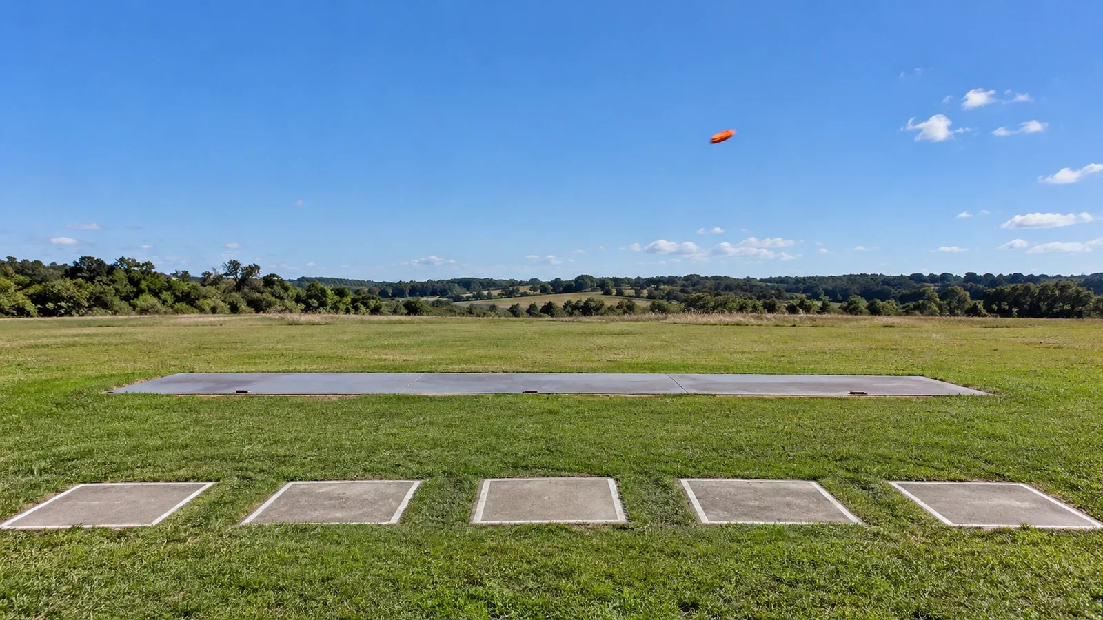 Campo de foso olímpico vacío, con cinco puestos de tiro, el foso de las máquinas y un plato naranja volando sobre el césped.